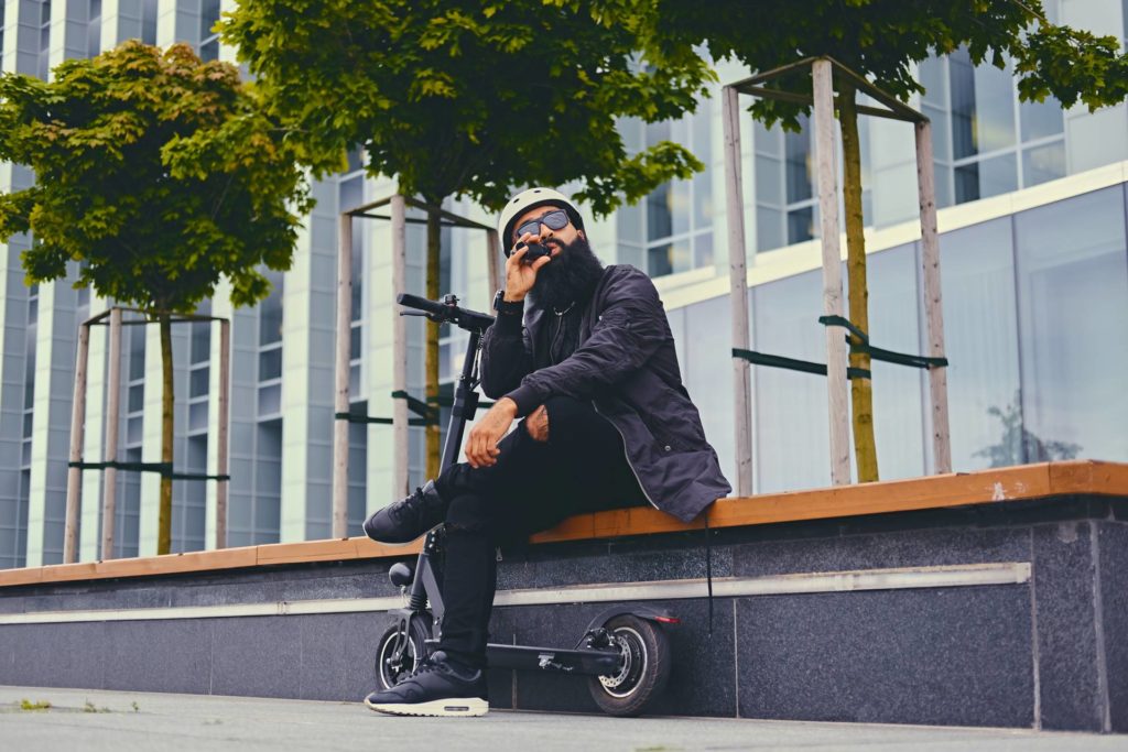 Ein bärtiger Hipster sitzt auf einer Bank neben seinem E-Scooter und dampft