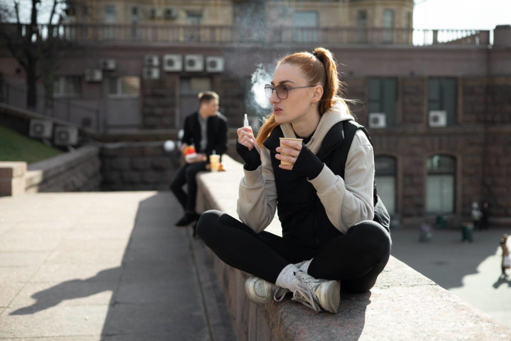 Junge Frau sitzt mit Kaffee und E-Zigaretteauf einer Mauer in der Stadt